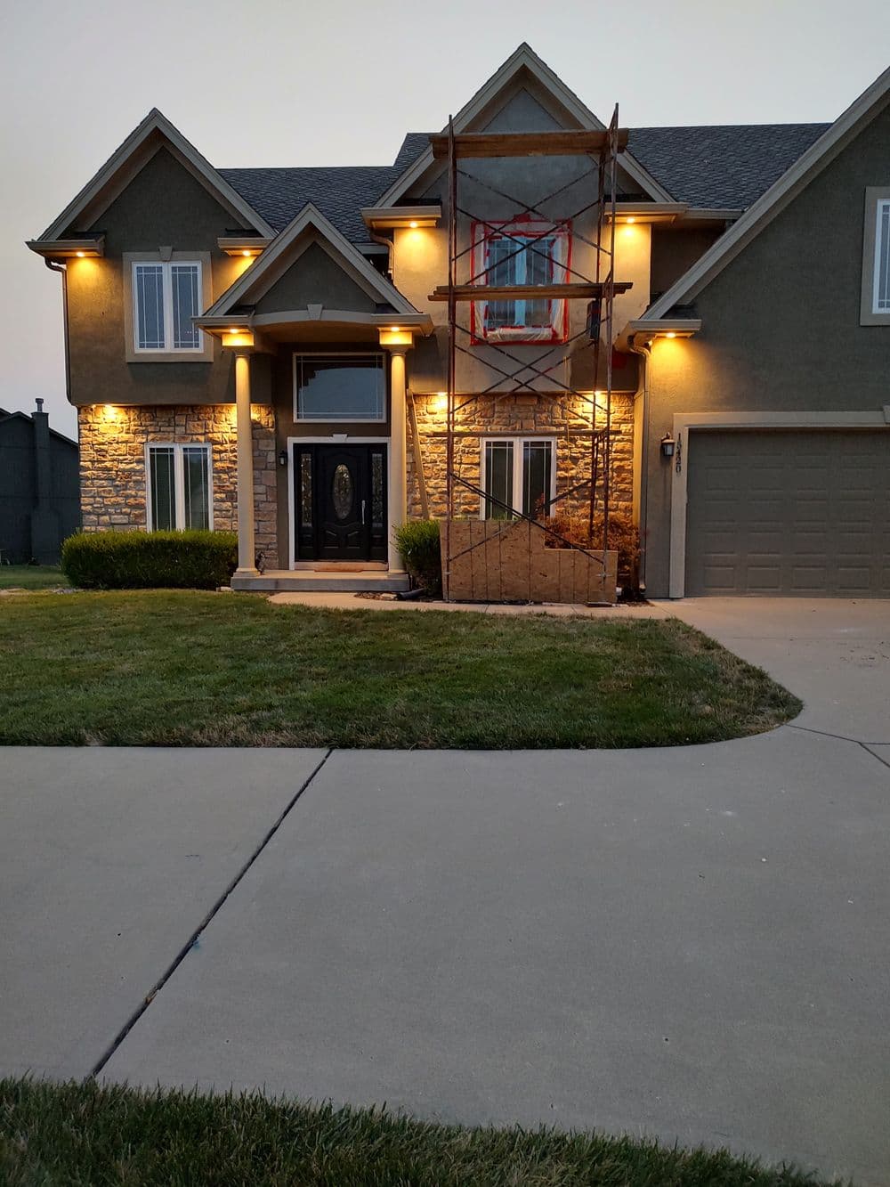 Two-story house with stone accents, scaffolding at the entrance, and glowing exterior lights.
